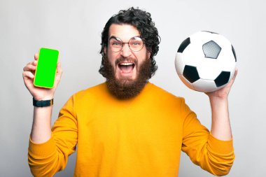 Sakallı bir adam kameraya çok heyecanlı bakıyor elinde bir telefon ve haykıran bir futbol topu var.