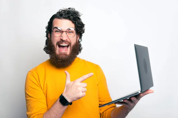 Aufgeregt blickt ein junger Mann in die Kamera und zeigt auf einen Computer, den er in der Hand hält. — Stockfoto