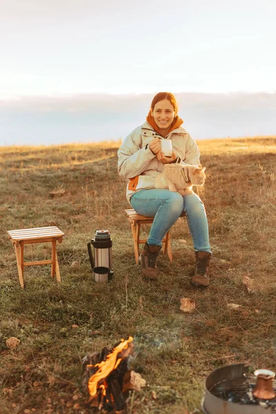 A smiling woman is sitting on a chair outside on a field drinking some tea — Stock Photo, Image