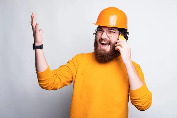 Egy fiatal, szemüveges szakállas építész beszél a telefonján egy fehér fal mellett. — Stock Fotó