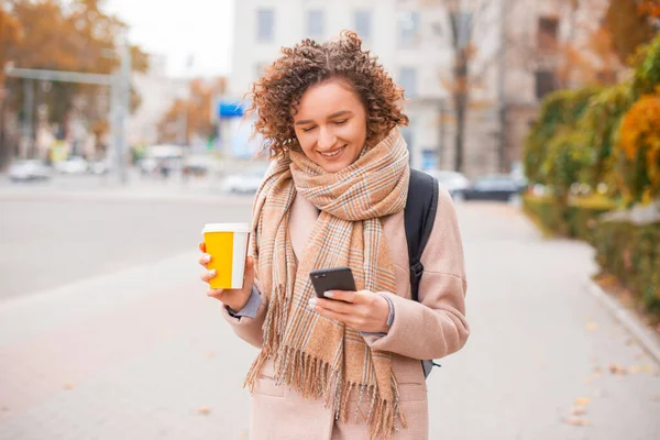 Uma mulher andando e segurando uma xícara de bebida quente está enviando mensagens de texto e sorrindo em seu telefone na cidade — Fotografia de Stock
