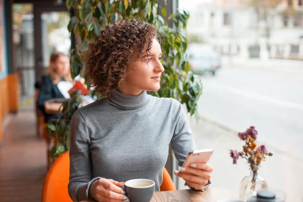 Uma jovem mulher está sittiong em uma mesa com uma xícara de café e um telefone está olhando para longe — Fotografia de Stock