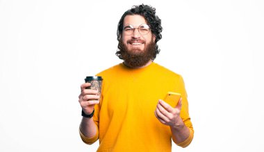 Yuvarlak gözlüklü mutlu sakallı adam kameraya bakıyor ve elinde kahve ve akıllı telefon tutuyor.