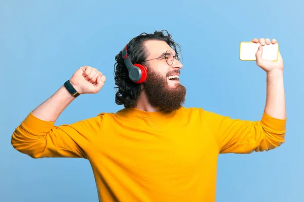 Alegre hombre barbudo guapo escuchando música en los auriculares y sosteniendo el teléfono inteligente —  Fotos de Stock