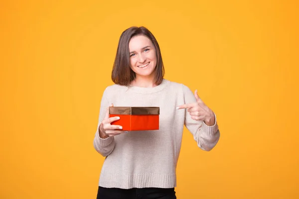 Veselá mladá žena ve svetru ukazuje na červený dárek přes žluté pozadí — Stock fotografie