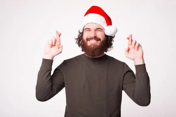 수염이 잘생긴 남자가 소원을 만들고 손가락을 교차하고 산타 클라우스 모자를 쓰고 있는 사진 — 스톡 사진