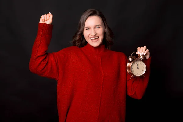 Spännande glad ung kvinna i röd tröja hålla klockan gör vinnaren gest på mörk bakgrund — Stockfoto