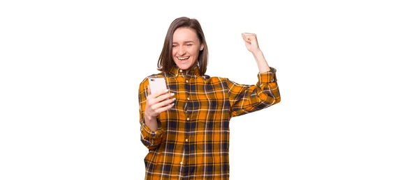 Retrato de uma menina bonita animado vestindo camisa xadrez olhando para o telefone celular e celebrando isolado sobre fundo branco — Fotografia de Stock