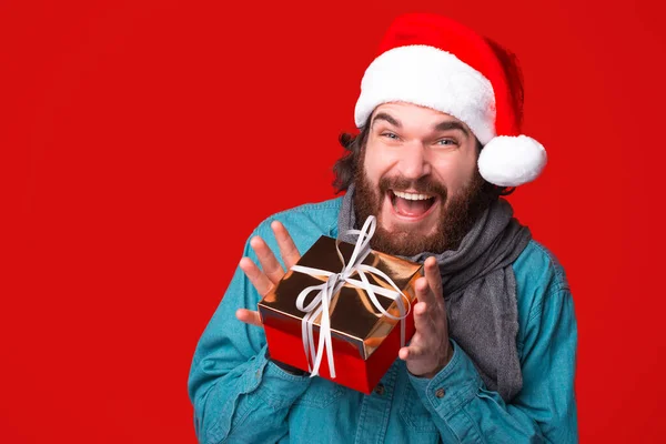 Podekscytowany brodaty mężczyzna z małym prezentem świątecznym, ubrany w czapkę Mikołaja, na czerwonym, odizolowanym tle — Zdjęcie stockowe