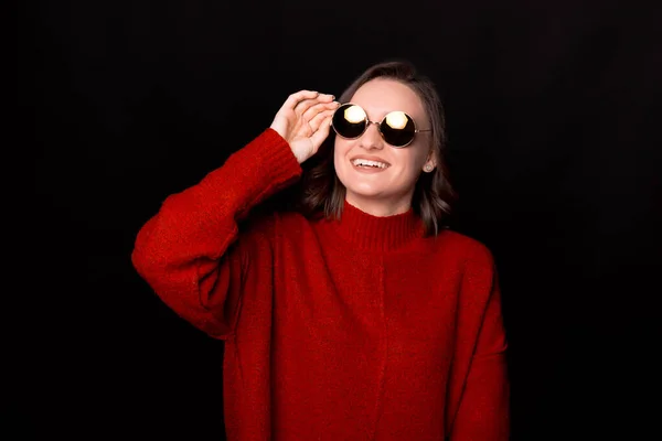 Fashionabla unga dam i röd tröja tittar bort och vidrör solglasögon. Inomhus porträtt av söt kvinna poserar på mörk bakgrund — Stockfoto