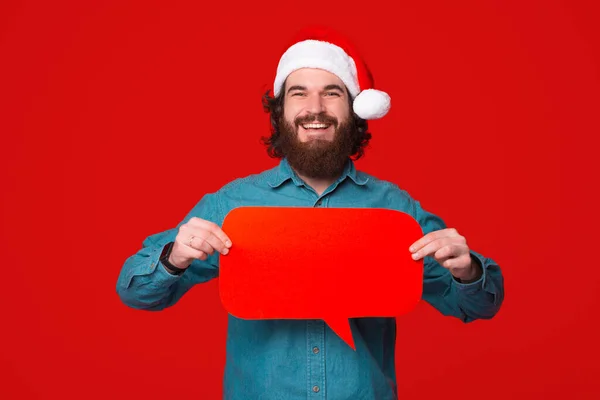 笑顔ひげを生やした男身に着けているクリスマスの帽子を保持していますスピーチバブル以上赤の背景 — ストック写真