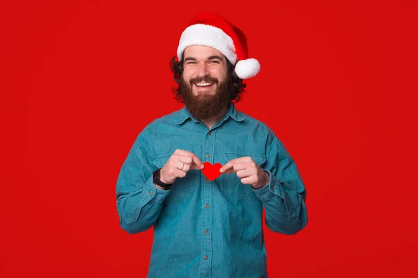 Χαρούμενος νεαρός που φοράει χριστουγεννιάτικο καπέλο κρατάει μια μικρή κόκκινη καρδιά — Φωτογραφία Αρχείου