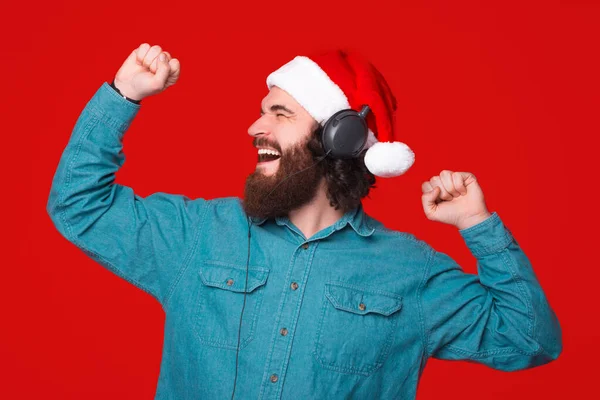 Podekscytowany młody brodaty mężczyzna w świątecznym kapeluszu słucha muzyki przez słuchawki i taniec. — Zdjęcie stockowe