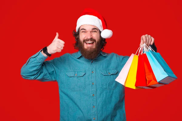 Jovem animado está segurando alguns sacos de compras e mostrando o polegar sobre o fundo vermelho. — Fotografia de Stock