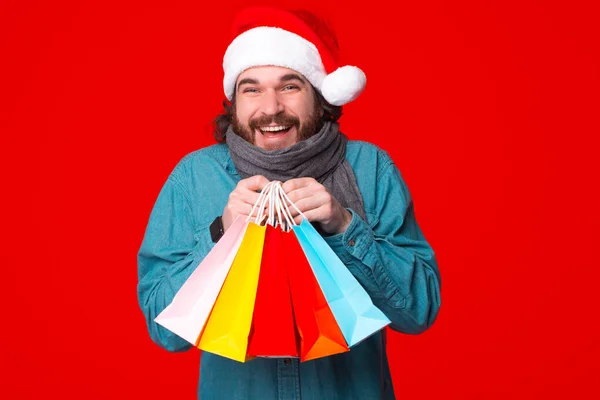 Święta Bożego Narodzenia. Facet trzyma torby na czerwonym tle. Hipster shopper szczęśliwy uśmiech w Santa kapelusz — Zdjęcie stockowe