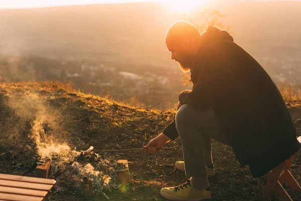 Photo of young man with beard is sitting near fire and cooking sausage on a field in the sunset — Stock Photo, Image