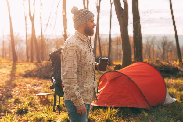 Foto av skäggig man med kopp kaffe, bär ryggsäck promenader nära sitt tält i skogen — Stockfoto