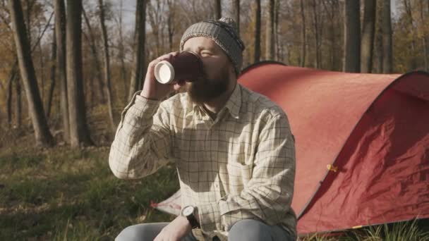 Footage of bearded hipster man sitting near tent and drinking his morning coffee — Stock Video