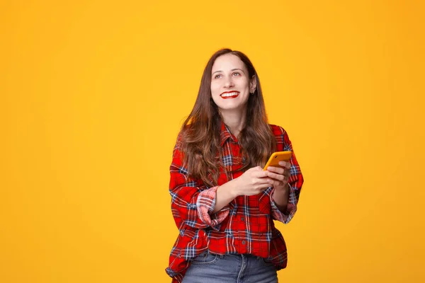 Foto de una joven alegre con camisa a cuadros sosteniendo el teléfono inteligente sonriendo y mirando hacia otro lado sobre el fondo amarillo — Foto de Stock