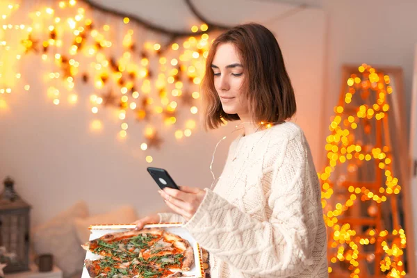 Jovencita sostiene un teléfono y una pizza en casa. — Foto de Stock