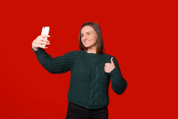 Menina sorrindo jovem está fazendo uma selfie com seu telefone sobre o fundo vermelho ao mostrar o polegar para cima. — Fotografia de Stock