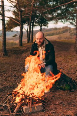 Sakallı adam kamp ateşinin yanında ısınıyor.