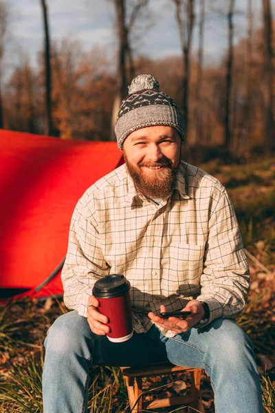Glad skäggig vandrare med smartphone och kopp i skogen — Stockfoto