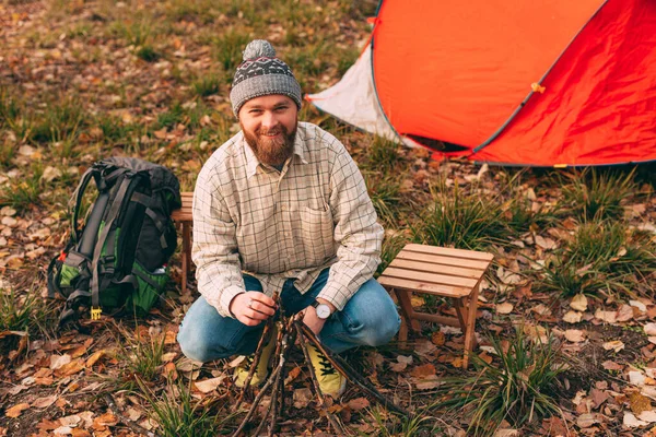 Positive bearded guy making fire near tent — Stock Photo, Image
