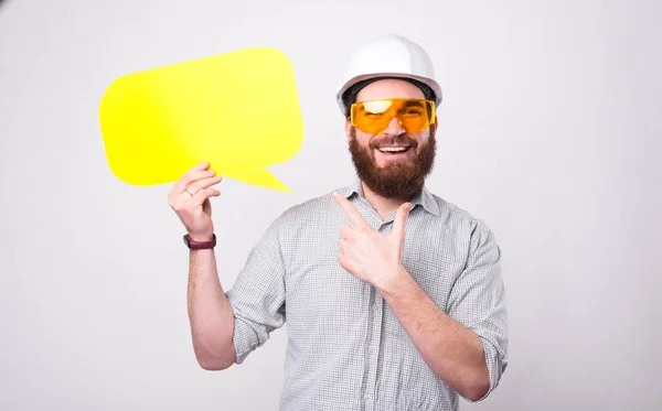 Vidám jóképű szakállas építész férfi védősisakban és rámutatva a sárga beszéd buborék — Stock Fotó