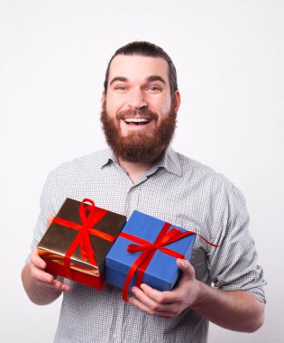 Neşeli, genç, sakallı bir adam kameraya bakıp mutlu görünüyor. Elinde beyaz bir duvarın yanında iki küçük hediye tutuyor.