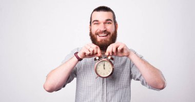 Yakışıklı, gülümseyen sakallı adam kameraya bakarak yüzünün yanında küçük sevimli bir saat tutuyor.