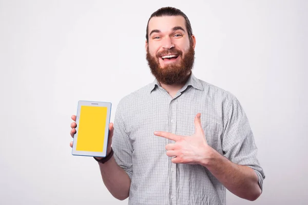 Jovem barbudo excitado está segurando um tablet e apontando para ele está sorrindo para a câmera perto de uma parede branca — Fotografia de Stock
