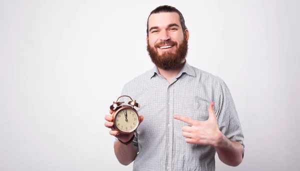 Un jeune homme positif sourit à la caméra et tient une horloge pointant vers elle près d'un mur blanc — Photo