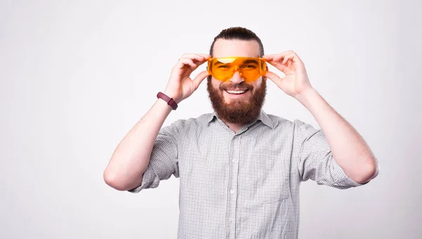 Boldog szakállas férfi silling a kamerába, és kezében egy pár védő szemüveg a szemét, közel a fehér fal — Stock Fotó