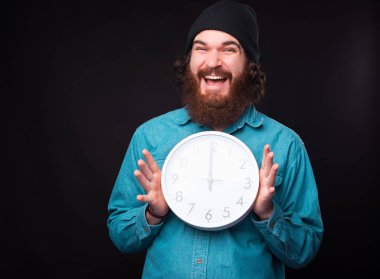 Neşeli genç bir adam kameraya gülümsüyor ve iki eli de siyah bir duvarın yanında yuvarlak beyaz bir saat tutuyor.