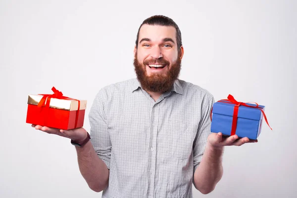 Un hombre barbudo feliz está mirando a la cámara y sostiene dos bonitos regalos tratando de ver cuál elegir. —  Fotos de Stock