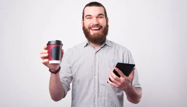 Un hombre barbudo alegre está sosteniendo su café y una tableta tomando su café sonríe a la cámara — Foto de Stock