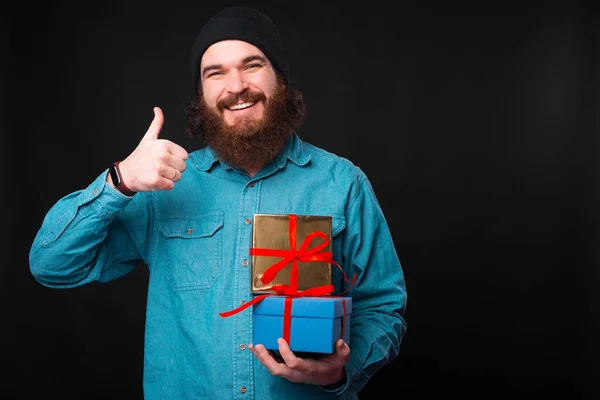 Szczęśliwy brodaty mężczyzna uśmiecha się do kamery i trzyma kciuk w górze, a niektóre prezenty pokazują, że lubi prezenty. — Zdjęcie stockowe