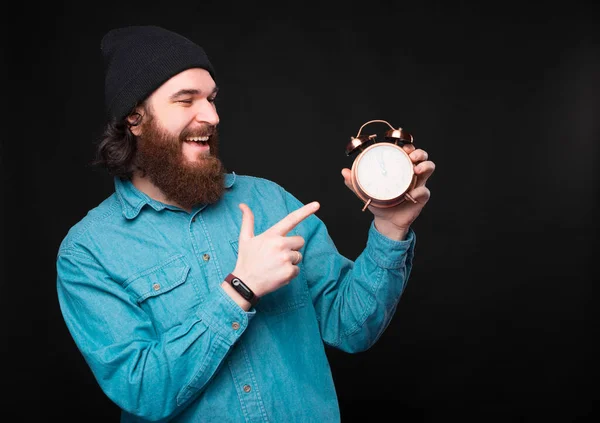 Ένας χαρούμενος νέος hipster κρατά ένα μικρό ρολόι και δείχνοντας σε αυτό χαμογελά δείχνοντας ότι είναι ικανοποιημένος με αυτό — Φωτογραφία Αρχείου