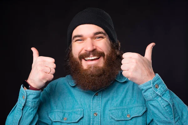 Ładny portret młodego brodatego hipstera uśmiechającego się do aparatu pokazującego obie krople w górę — Zdjęcie stockowe