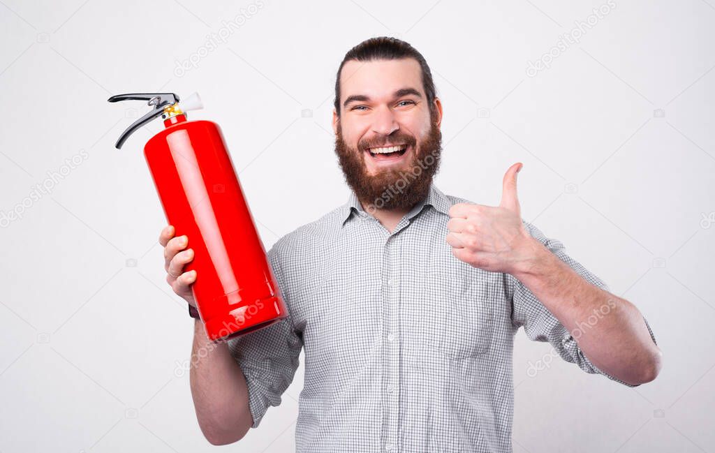 A young bearded man is looking and smiling at the camera holding a fire extinguisher and a thumg up near a white wall