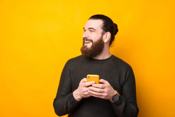 Joyful bearded hipster man standing over yellow background using a smartphone and looking away at copyspace — Stock Photo, Image