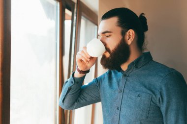 Sakallı bir ofis çalışanının pencerede oturup kahve içerkenki fotoğrafı.