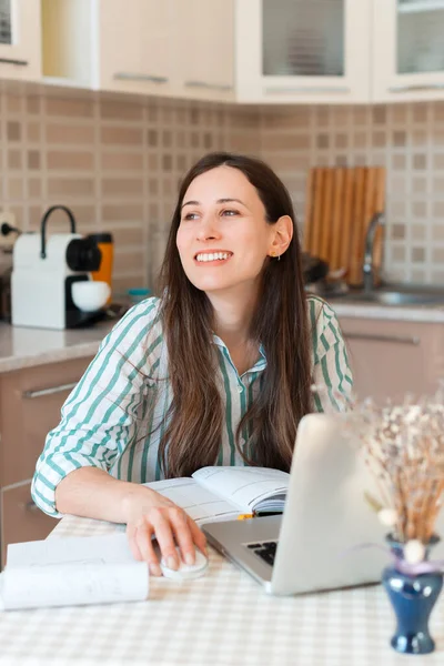 Porträtt av leende ung kvinna och tittar bort medan du sitter vid skrivbordet i köket — Stockfoto
