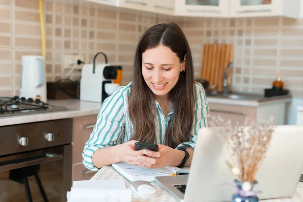 Joven alegre enviando un mensaje de texto en el teléfono inteligente mientras trabaja desde casa en la cocina — Foto de Stock