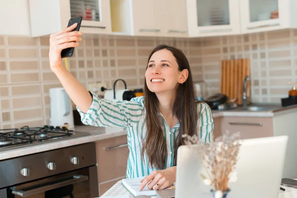 Jovencita alegre tomando selfie en la cocina mientras trabaja desde casa en el portátil — Foto de Stock