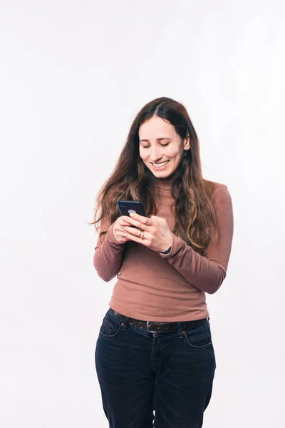 Cheerful young woman texting message on smartphone over white wall — Stock Photo, Image