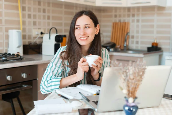 Счастливая молодая женщина держит чашку кофе и сидит перед ноутбуком, работает из дома — стоковое фото