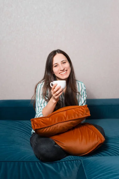 Jovem encantadora sentada no sofá e segurando xícara de café — Fotografia de Stock