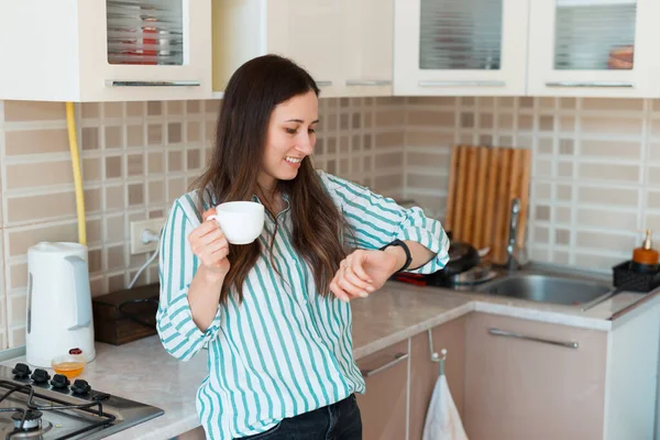 Foto de una mujer joven en posición informal en la cocina con taza de café y mirando smartwatch — Foto de Stock